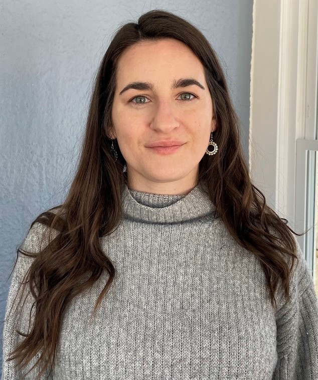 Alison Cameron with long dark hair, green eyes, light skin; wearing a gray sweater and earrings; in front of a blue wall. 
